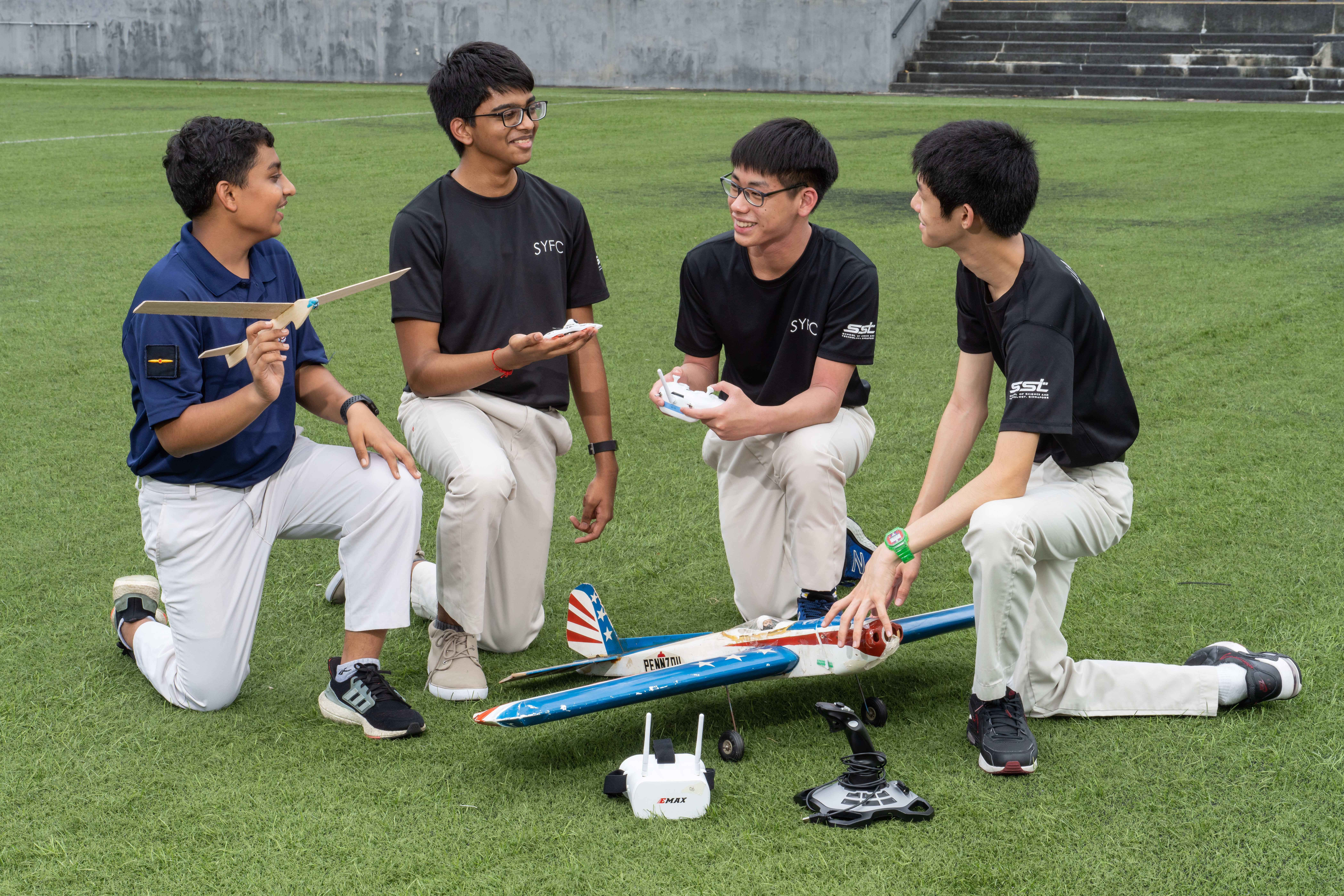 Four teens kneel on grass around a model airplane and remote controls, one holds the plane's wing.